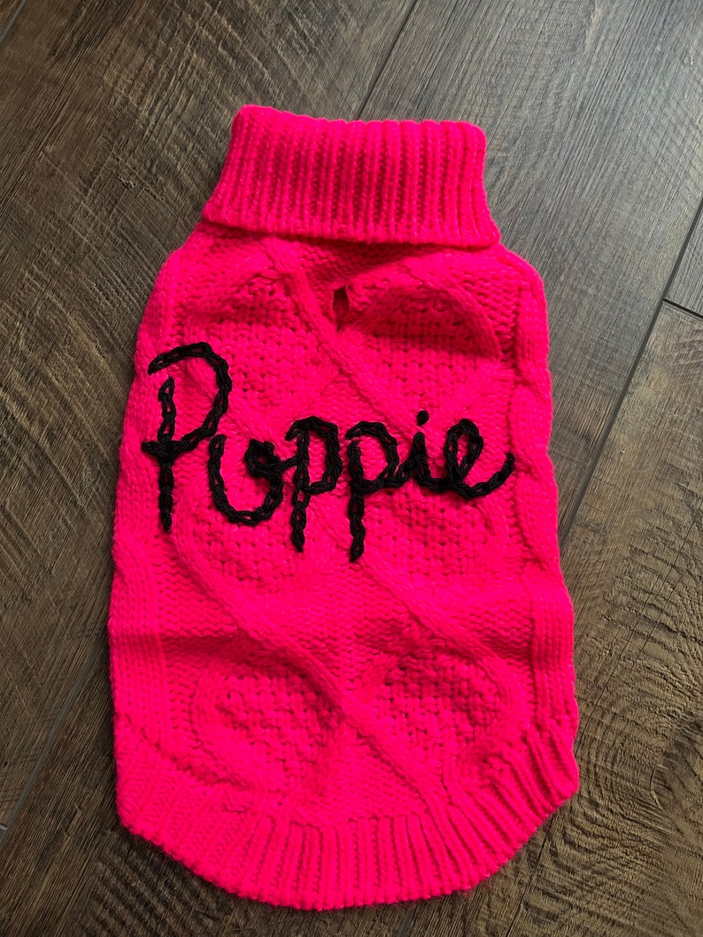 K&ouml;nnte beinhalten: Ein rosa gestrickter Hundepullover mit einem schwarzen Zopfmuster und dem Namen "Poppy" auf der Vorderseite gestickt.