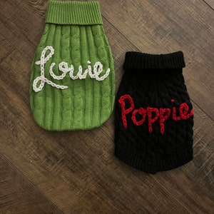 K&ouml;nnte beinhalten: Zwei gestrickte Hunde-Pullover, einer gr&uuml;n mit wei&szlig; gestickten Buchstaben, die "Louie" sagen, und einer schwarz mit rot gestickten Buchstaben, die "Poppy" sagen.