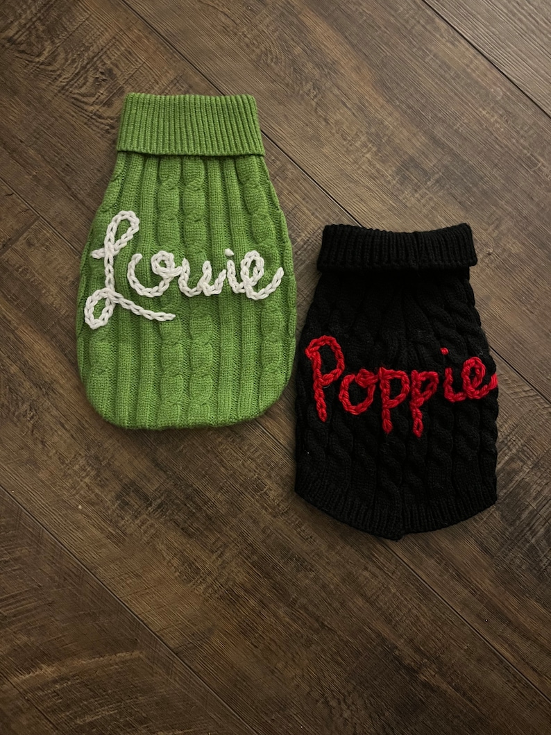 K&ouml;nnte beinhalten: Zwei gestrickte Hunde-Pullover, einer gr&uuml;n mit wei&szlig; gestickten Buchstaben, die "Louie" sagen, und einer schwarz mit rot gestickten Buchstaben, die "Poppy" sagen.