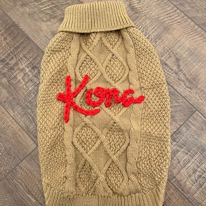 K&ouml;nnte beinhalten: Ein beiger Strickpullover mit Zopfmuster f&uuml;r Hunde mit dem roten Stickwort "Kona" auf der Vorderseite.