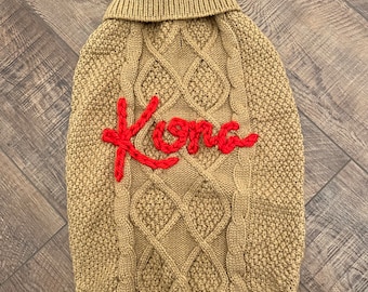 May include: A tan cable knit dog sweater with a red embroidered word "Kona" on the front.