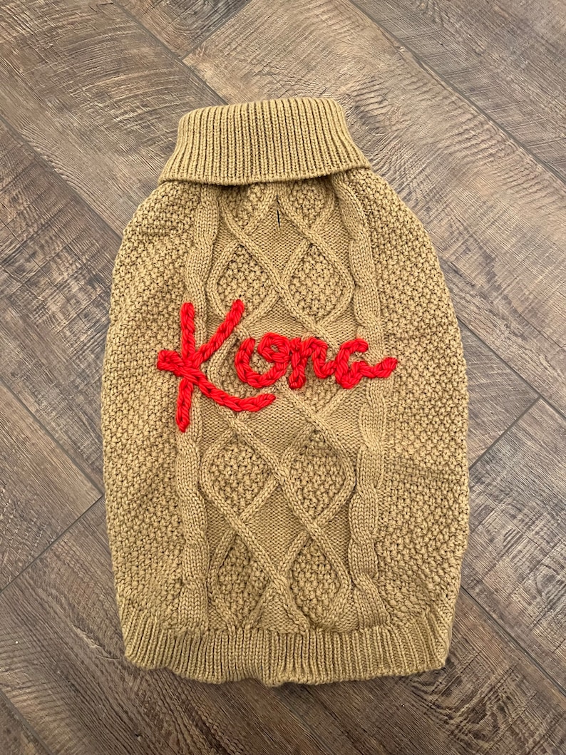 K&ouml;nnte beinhalten: Ein beiger Strickpullover mit Zopfmuster f&uuml;r Hunde mit dem roten Stickwort "Kona" auf der Vorderseite.