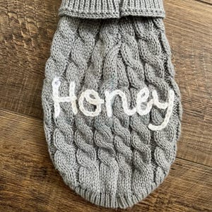 K&ouml;nnte beinhalten: Grauer Strickpullover f&uuml;r Hunde mit einem wei&szlig;en gestickten Namen "Honey".