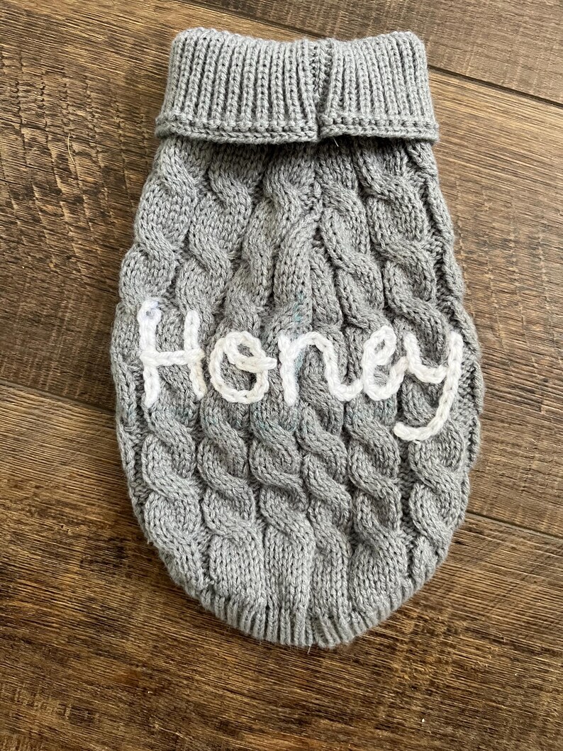 K&ouml;nnte beinhalten: Grauer Strickpullover f&uuml;r Hunde mit einem wei&szlig;en gestickten Namen "Honey".