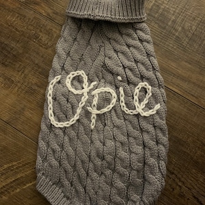 K&ouml;nnte beinhalten: Grauer Strickpullover f&uuml;r Hunde mit Zopfmuster und dem Namen "Opie" in wei&szlig; gestickt.