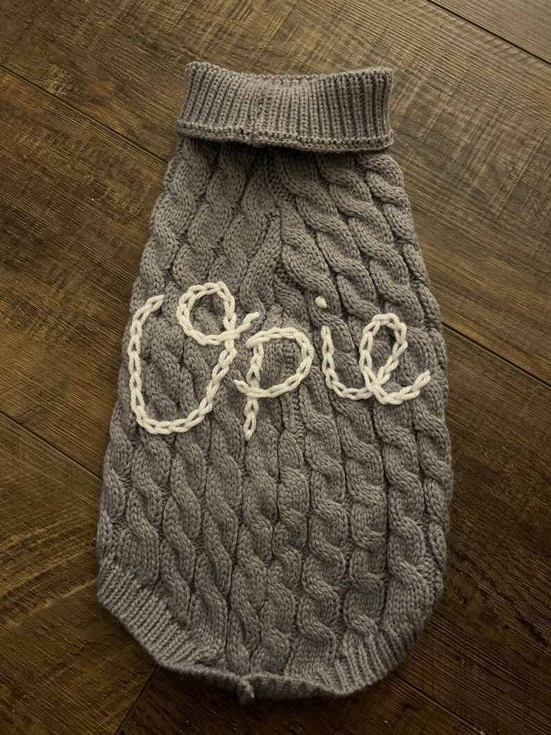 K&ouml;nnte beinhalten: Grauer Strickpullover f&uuml;r Hunde mit Zopfmuster und dem Namen "Opie" in wei&szlig; gestickt.