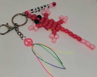 May include: A handmade beaded keychain featuring a pink lizard, a peace sign charm with colorful strands, and a beaded name tag that says "JENNY". The lizard is made of pink and black beads.