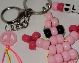 May include: A handmade keychain featuring a pink peace sign charm, a beaded figure, and a beaded word. The beaded figure is made of pink beads with black accents. The keychain also includes a silver keyring and lobster clasp.