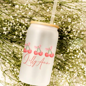 May include: A white glass can-shaped tumbler with a bamboo lid and a clear straw. The tumbler features a design of three sets of pink cherries with pink bows and the name "Lilly Ann" in pink script. The background is white with small white flowers.