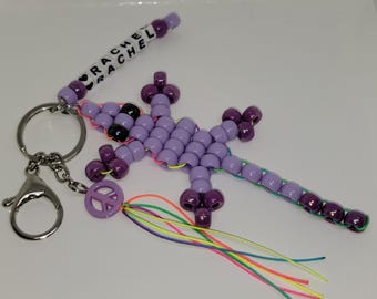 May include: A handmade beaded keychain featuring a purple lizard design. The keychain includes a silver clasp, a peace sign charm, and a beaded section with the name "RACHEL". The lizard has black and purple beads with a colorful tassel.