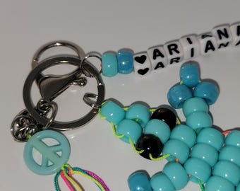 May include: A handmade keychain featuring a peace sign charm, a beaded design with the name "ARIANA", and a collection of blue and black beads. The keychain includes a silver-colored metal ring and clasp.