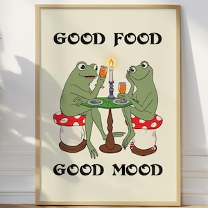 Puede incluir: Una impresión artística enmarcada con dos ranas verdes de dibujos animados en una mesa, disfrutando de bebidas. Están sentadas en taburetes de seta, con una vela y las palabras "GOOD FOOD GOOD MOOD" en texto negro. Estilo retro y caprichoso.
