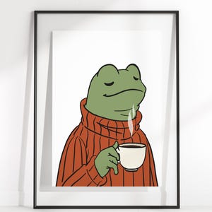 Puede incluir: Impresión artística enmarcada que presenta una rana de dibujos animados con un suéter color óxido y una taza de café blanca. La rana tiene los ojos cerrados y sale vapor del café. La obra de arte está sobre un fondo blanco.