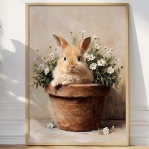 Puede incluir: Una impresión artística enmarcada que presenta un conejo marrón claro en una maceta de terracota llena de flores blancas y follaje verde. El conejo tiene orejas largas y un pelaje esponjoso. La obra de arte está en un marco dorado.