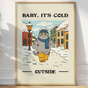 Puede incluir: Un estampado enmarcado con el texto "BABY, IT'S COLD OUTSIDE". La ilustración presenta un gato gris con bufanda azul, gorro y botas amarillas, caminando por una calle nevada con edificios coloridos y una farola.