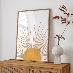Puede incluir: Una impresión enmarcada de un sol estilizado con un centro amarillo y rayos marrones. La impresión está colgada en una pared sobre un armario de madera con un frente de ratán tejido.