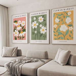 Puede incluir: Tres impresiones florales enmarcadas con el texto "FLOWER MARKET" y el nombre de la galería de arte en la parte inferior. Las impresiones presentan flores blancas estilizadas con hojas verdes sobre un fondo rosa, verde y amarillo.