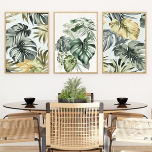 Puede incluir: Tres impresiones enmarcadas con ilustraciones de acuarela de hojas tropicales en tonos verdes y amarillos. Las impresiones están colgadas en una pared blanca sobre una mesa de comedor de madera con cuatro sillas.