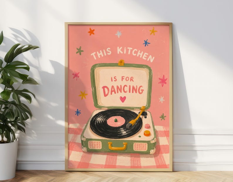 May include: A framed art print featuring a vintage-style record player inside a green suitcase, set against a pink background with the text "THIS KITCHEN IS FOR DANCING." Colorful stars and a heart accent the design. The print is in a wooden frame.