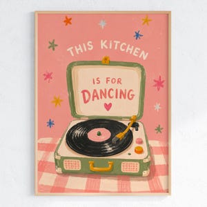 May include: A framed art print featuring a vintage-style record player with the text "THIS KITCHEN IS FOR DANCING" on a pink background. The record player is green and white, with a black record and a yellow tone arm. Colorful star shapes are scattered around the text.