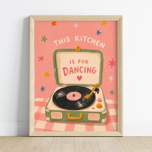 May include: A framed art print featuring a vintage-style record player with the text "THIS KITCHEN IS FOR DANCING." The artwork has a pink background with colorful star accents and a checkered pattern at the bottom.