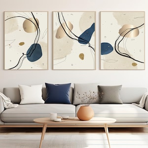 Puede incluir: Tres impresiones de arte abstracto con fondo blanco y formas abstractas doradas y azules. Las impresiones están colgadas en una pared blanca sobre un sofá gris claro con almohadas.
