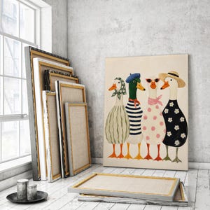 May include: A canvas print featuring four whimsical ducks, each adorned in unique outfits: a green scarf, a blue beret, sunglasses, and a straw hat. The ducks stand in a row against a neutral background. Several blank canvases lean against a wall.