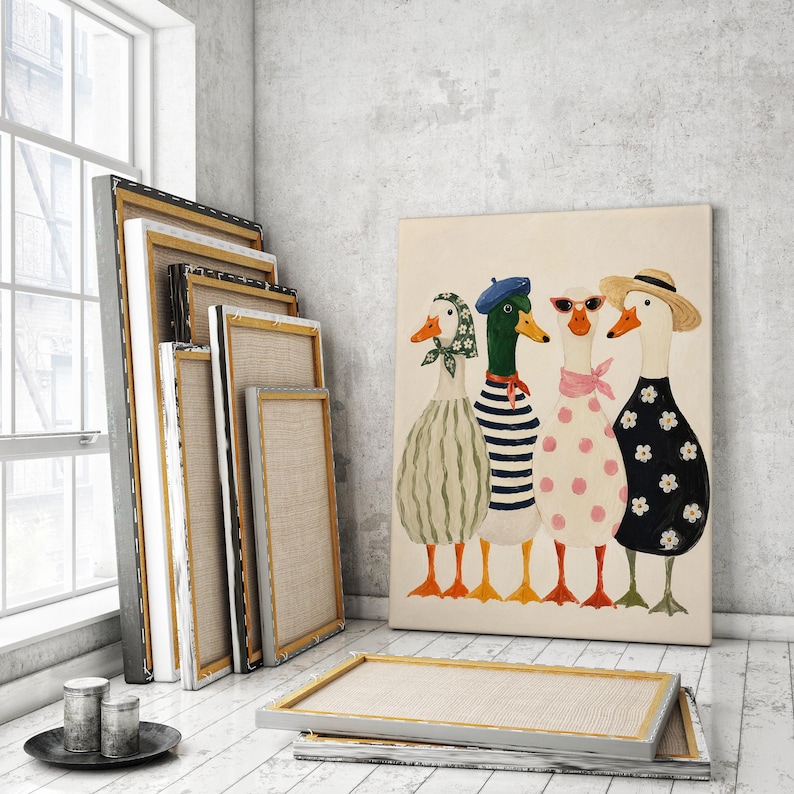 May include: A canvas print featuring four whimsical ducks, each adorned in unique outfits: a green scarf, a blue beret, sunglasses, and a straw hat. The ducks stand in a row against a neutral background. Several blank canvases lean against a wall.