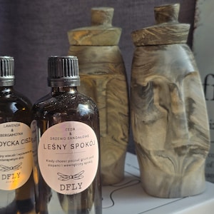 May include: An assortment of brown glass bottles with black caps and labels, alongside decorative stone-like jars. The bottles have labels with text in Polish. The jars are in shades of gray and beige.