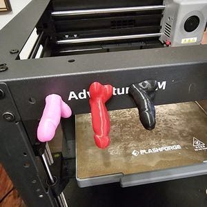 Puede incluir: Una impresora 3D con un marco negro y una cama de impresión gris. La cama de impresión tiene el texto "FLASHFORGE" impreso en ella. Hay tres objetos impresos en 3D en la impresora: un objeto rosa, un objeto rojo y un objeto negro. Los objetos tienen todos la forma de corazones.