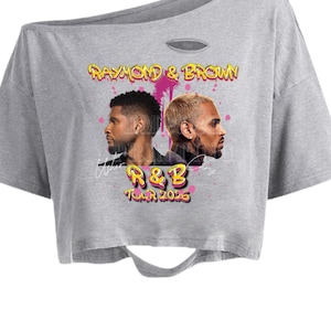 Puede incluir: Top corto gris con el texto "RAYMOND & BROWN" y "R&B TOUR 2026" en amarillo y rosa. La camiseta tiene un dise&ntilde;o desgastado con dos retratos masculinos. La parte superior tiene un escote asim&eacute;trico.