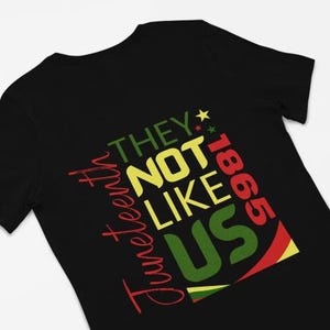 Può includere: T-shirt nera con la scritta "Juneteenth" in rosso, "THEY NOT LIKE US" in giallo e verde e "1865" in rosso. Il design incorpora i colori della bandiera panafricana.