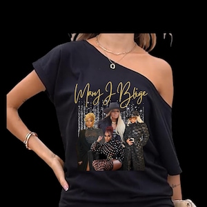 May include: Black off-the-shoulder top featuring a graphic of Mary J. Blige and other women. The text "Mary J. Blige" is in gold script. The graphic includes images of women in various outfits, set against a sparkling background.