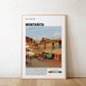 May include: A framed print featuring the word "MONTANITA" in bold black letters, with "ECUADOR" above it. The image shows a street scene with colorful buildings, thatched roofs, and people. The print has a light-colored wooden frame.
