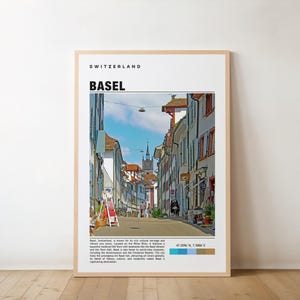 May include: A framed print featuring a street scene in Basel, Switzerland. The image shows a narrow street lined with buildings, a church steeple in the distance, and the text "Basel" and "Switzerland" at the top. The print is in a light-colored frame.