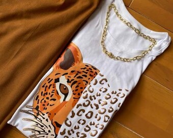 May include: A white t-shirt with a gold chain necklace and a large leopard graphic. The shirt is paired with brown pleated trousers. The leopard design features orange, brown, and white colors. The outfit is displayed on a wooden surface.
