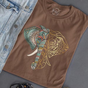 May include: A brown t-shirt featuring a detailed elephant design with gold and colorful accents. The shirt is paired with a light blue denim jacket, sunglasses, and a beaded bracelet. The brand logo "K&L" is visible on the shirt.