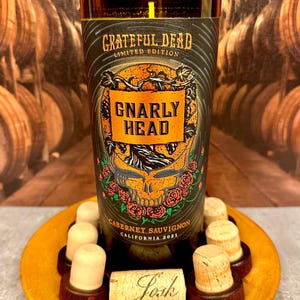 May include: A candle in a repurposed wine bottle with a label that reads "Grateful Dead Limited Edition Gnarly Head Cabernet Sauvignon California 2021." The candle sits on a wooden plate with wine corks.