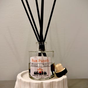 May include: A clear glass reed diffuser labeled "Rum Punch" with black reeds, a wooden stopper, and a white tree stump-shaped base. The label reads "HORN BUILT CANDLES".