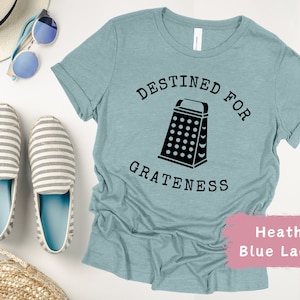 May include: A light blue t-shirt with the text "Destined for Grateness" and a black graphic of a grater. The text is in a black typewriter font. The t-shirt is on a white background with a straw hat, sunglasses, and striped shoes.