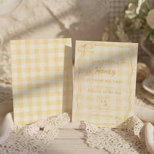 May include: Two rectangular cards with a yellow and white gingham pattern. The right card features yellow line art of ribbons, bees, and text reading Our Little Honey is turning one! Natalia's 1st Bee-Day, with date and location details.