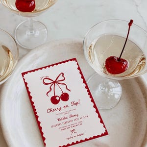 Op de afbeelding: Een rechthoekige uitnodigingskaart met een geschulpte rode rand, voorzien van een rode illustratie van twee kersen met een strik. De tekst luidt: she's our Cherry on Top! Please join us for a baby shower honoring Natalia Gomez, Saturday February 28th at 3 PM, 214 Cherry Ln, Bow City 29408, kindly RSVP to Mia by 2.20, registry on babylist.