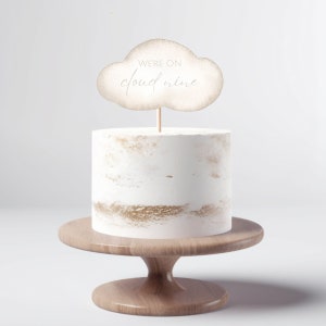 Puede incluir: Un pastel blanco con detalles dorados sobre un soporte de pastel de madera. Un adorno de pastel en forma de nube con el texto "We're on cloud nine" está en la parte superior del pastel.