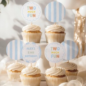 Op de afbeelding: Zes cupcakes met witte glazuur, elk voorzien van een ronde papieren topper op een houten prikker. De toppers hebben lichtblauwe strepen of geschulpte randen met de tekst "TWO MUCH FUN" of "ALEX'S 2ND BIRTHDAY" in blauw, geel en oranje.