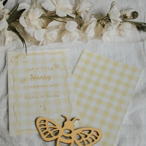 May include: A yellow and white gingham birthday invitation card with the text Our Little Honey is turning one, Natalia's 1st Bee-Day, Aug 20, and a wooden bee cutout. The invitation features line art of bees and ribbons on a checkered background.