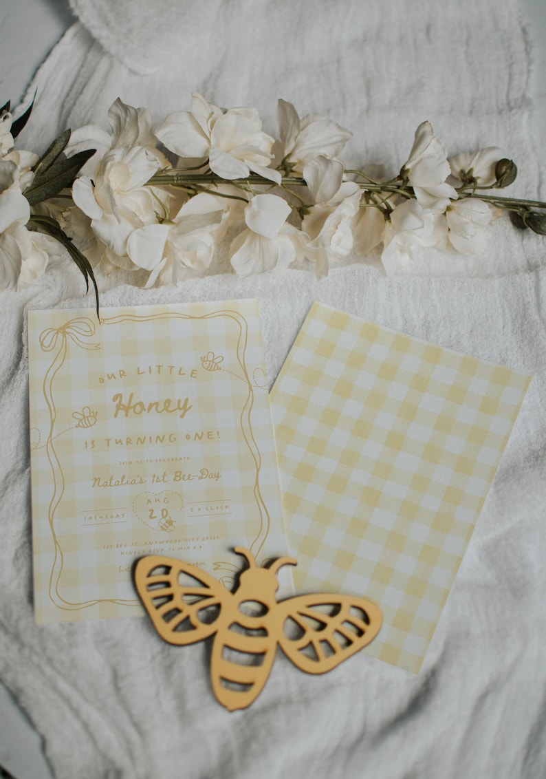 May include: A yellow and white gingham birthday invitation card with the text Our Little Honey is turning one, Natalia's 1st Bee-Day, Aug 20, and a wooden bee cutout. The invitation features line art of bees and ribbons on a checkered background.