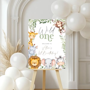 Può includere: Un cartello di benvenuto verde e bianco con il testo "Wild One" e "Benvenuto al 1° compleanno di Alex!" circondato da illustrazioni di animali della giungla, tra cui una giraffa, un rinoceronte, un leone, un elefante, una zebra e una scimmia.