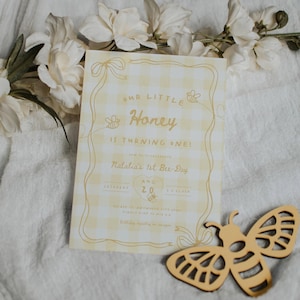 May include: A rectangular birthday invitation card with a yellow and white gingham pattern, featuring the text Our Little Honey Is Turning One, Natalia's 1st Bee-Day, Aug 20, and bee illustrations, accompanied by a wooden bee cutout.