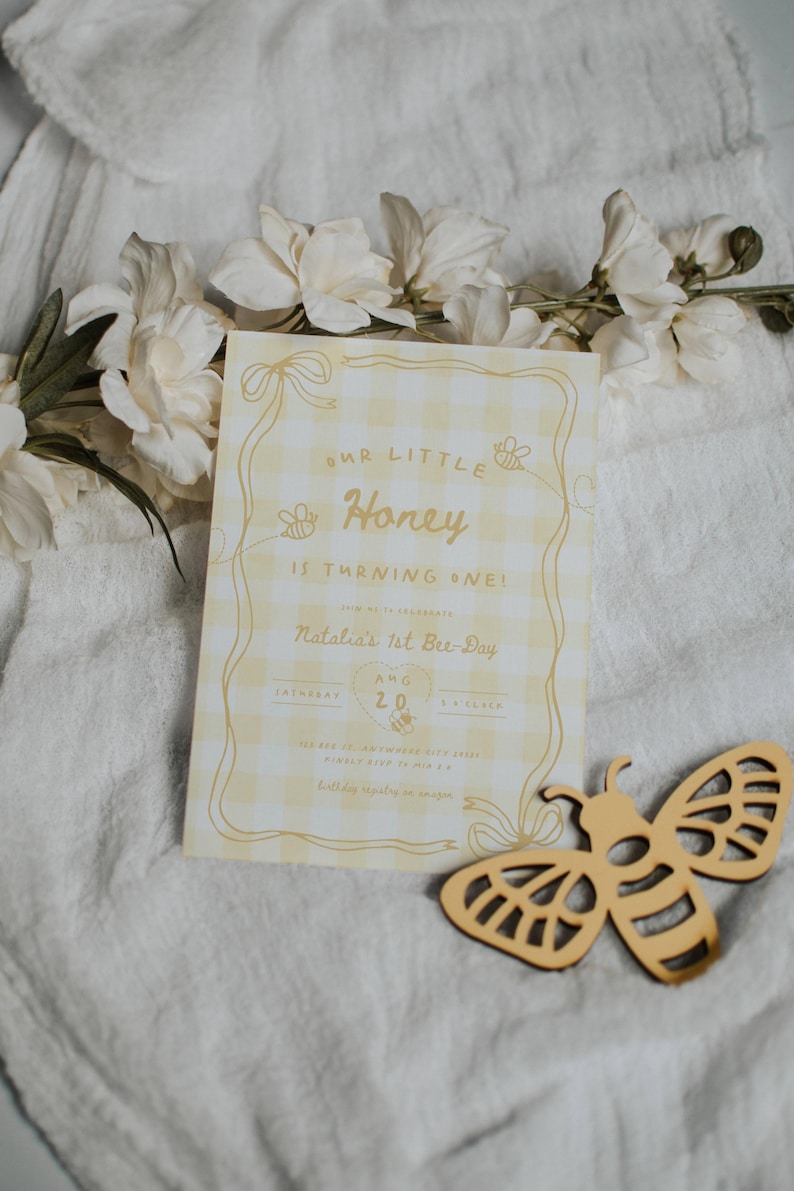 May include: A rectangular birthday invitation card with a yellow and white gingham pattern, featuring the text Our Little Honey Is Turning One, Natalia's 1st Bee-Day, Aug 20, and bee illustrations, accompanied by a wooden bee cutout.