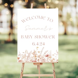 May include: A white sign on a wooden easel with the words "WELCOME TO Summer's BABY SHOWER 6.4.24" in a soft brown font. The bottom of the sign features a watercolor floral design in shades of peach, tan, and brown.